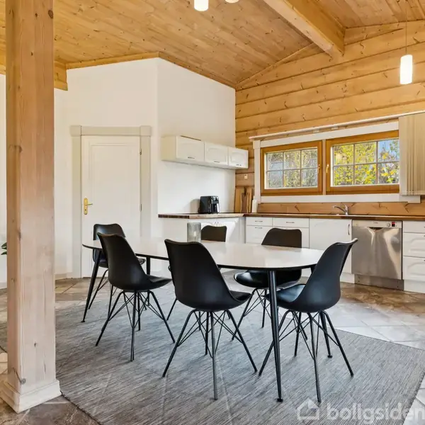 Et spisebord med seks sorte stole står på et tæppe i et køkken med træmøbler. Vinduet i baggrunden giver udsigt til natur.