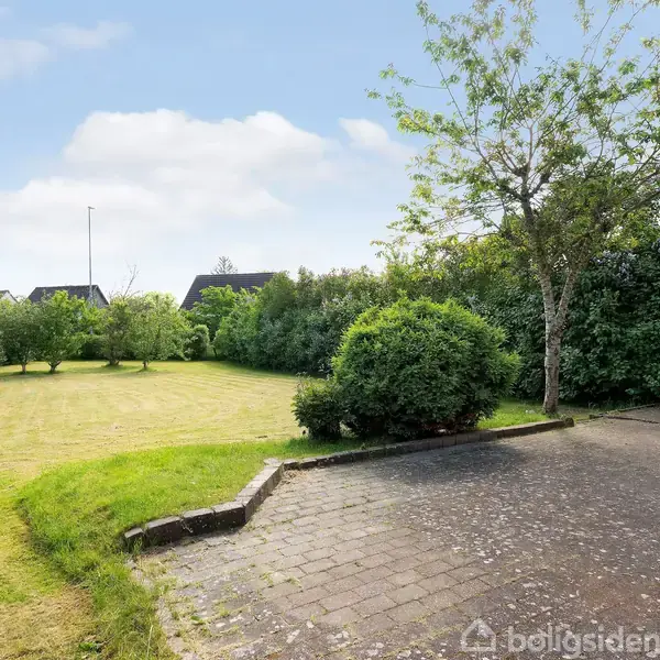 En velplejet græsplæne med omgivende hække og træer i en boligområde. Til højre ses en flisebelagt indkørsel. Skyer pryder en blå himmel.