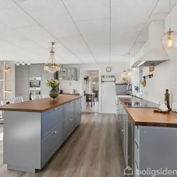 Køkkenet har en lang ø med grå skabe og træbordplade, omgivet af hvide vægge og store vinduer. Der er også moderne apparater og hængelamper, der skaber en lys og åben atmosfære.