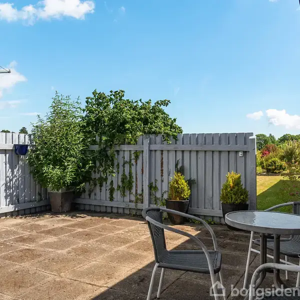 Et rundt bord med to stole står på en flisebelagt terrasse. I baggrunden ses en grå træhegn og grønne planter. Det er solrigt med blå himmel og få skyer.