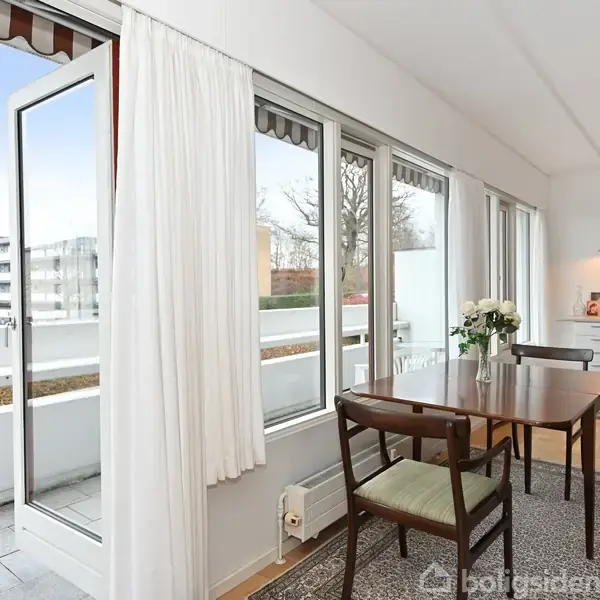 Spisestue med træbord og stole står ved store vinduer mod en åben terrasse. I baggrunden ses et skrivebord med lampe og billeder. En lys og moderne indretning.