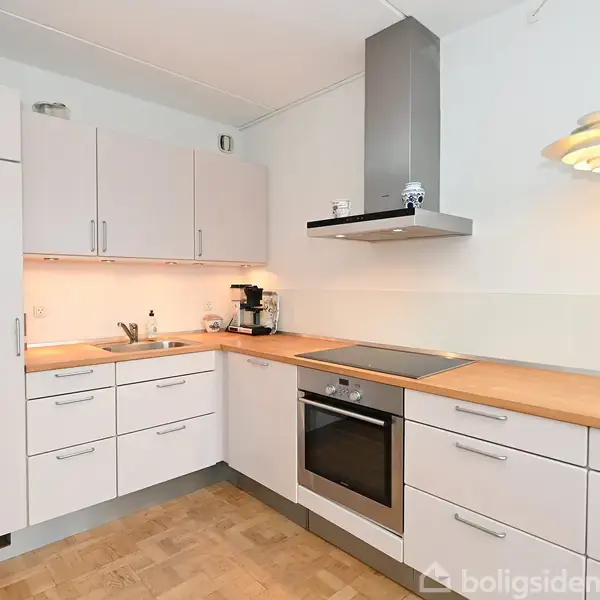 Et moderne køkken med hvide skabe, en indbygget ovn og kogeplade. En kaffemaskine står på bordpladen. Rummet har lyse vægge og træparketgulv.