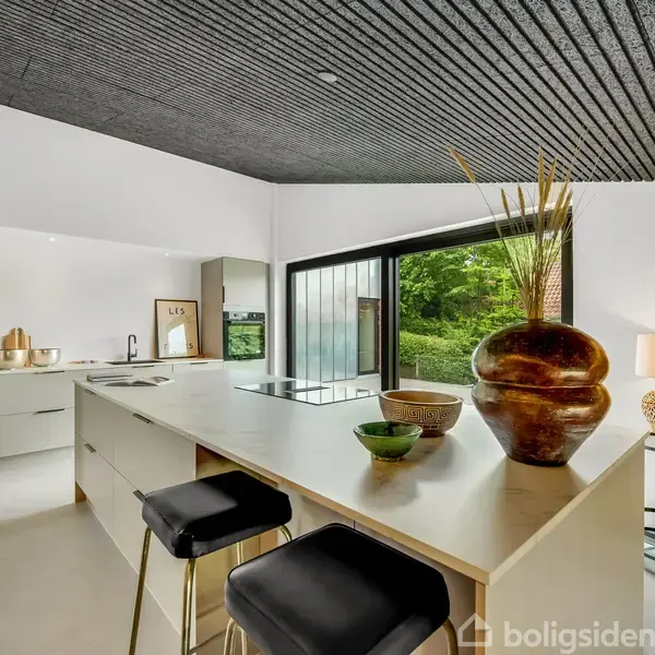 Køkken med en moderne ø og barstole, dekoreret med store keramikvaser og skåle. Store vinduer giver udsigt til en grøn have. Sort loft og hvidmalede vægge.