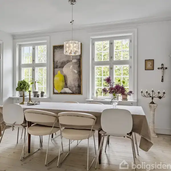 Et spisebord står dækket med en dug og omgivet af stole, placeret foran to store vinduer. En lysekrone hænger over bordet. Et skab og dekorationer fuldfører rummet.