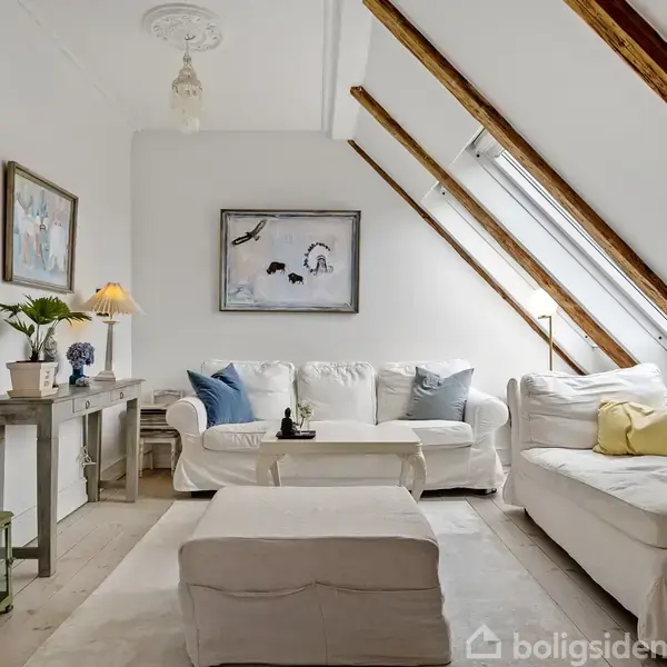 En lys stue med hvid sofa og skammel, pyntet med farverige puder. Skråvinduer slipper sollys ind. Et lille bord og konsolbord med lamper og planter supplerer rummet.
