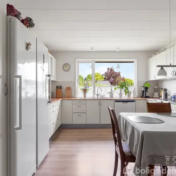Moderne køkken med hvidt interiør, spisebord med dug og stole i forgrunden. Rummet er lyst med trælaminatgulv, store vinduer og blomster på bordet. En pendellampe hænger over bordet.