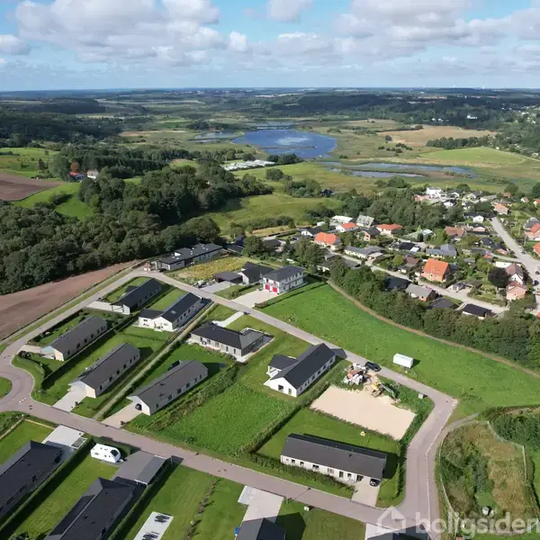 Et boligområde med rækkehuse omgivet af grønne marker ses fra luften. Der er en rundkørsel tæt på husene og en sø i baggrunden under en blå himmel.