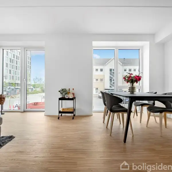 Stuen indeholder et rundt spisebord med sorte stole i et lyst rum med trægulv. Store vinduer giver udsigt til en moderne bygning udenfor. En sofa og planter kompletterer indretningen.