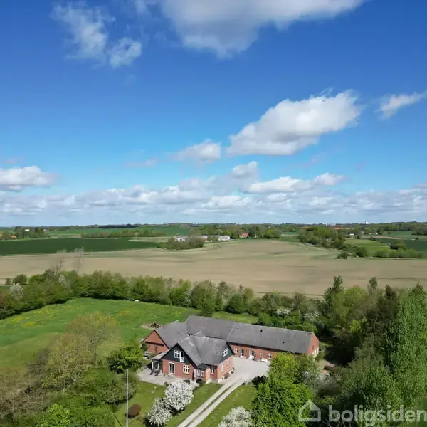 En stor ejendom ligger i et landligt område omgivet af åbne marker og grønne træer under en blå himmel med få skyer.