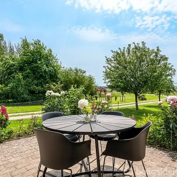 Rundt havebord med fire stole står på en stenbelagt terrasse. Omgivet af blomster og buske; grønt græs og træer i baggrunden. Et dansk flag vajer til højre.
