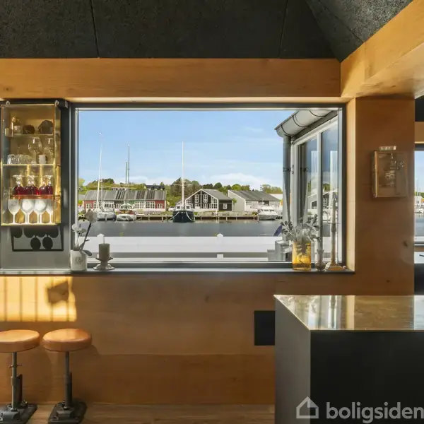 Barstole står stille ved en træbar, mens store vinduer viser en marina fuld af både i baggrunden. Interiøret er moderne med en minimalistisk stil.