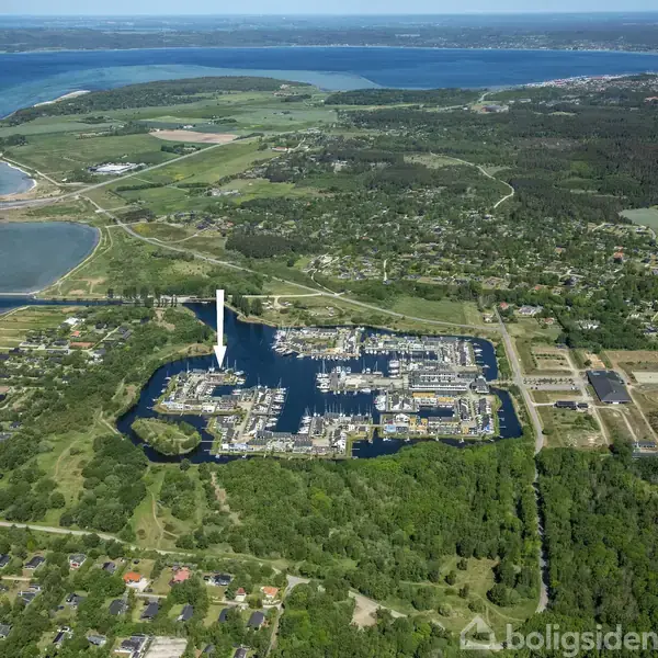 Luftfoto af marina med både i vandet; vindmøller i baggrunden. Omkringliggende landskab viser grønne skove, marker og en kystlinje med åbent hav i horisonten.