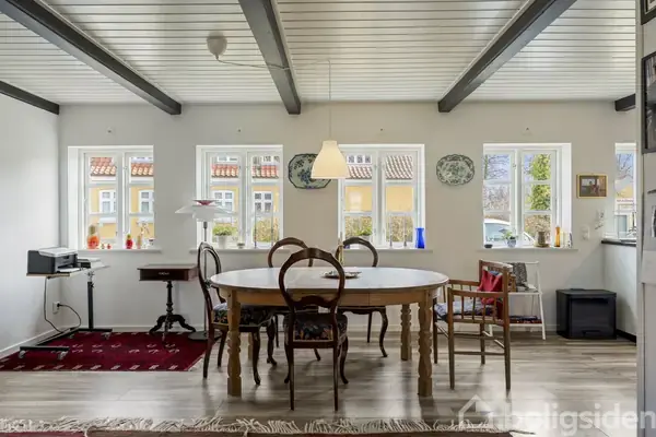 Et rundt spisebord med stole står i et lyst alrum. Vinduessæt med fire ruder og dekorationer i baggrunden. Træbjælker ses i loftet, og gulvet er af træplanker.
