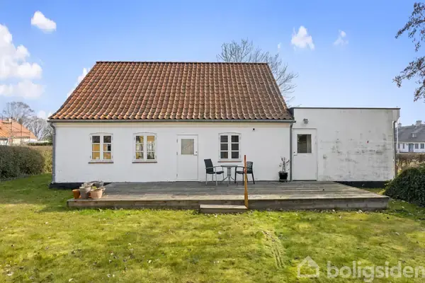 Hvidt hus med rødt tegltag står stille i et solbeskinnet haveområde. Foran huset er en træterrasse med to sorte stole ved et lille bord.