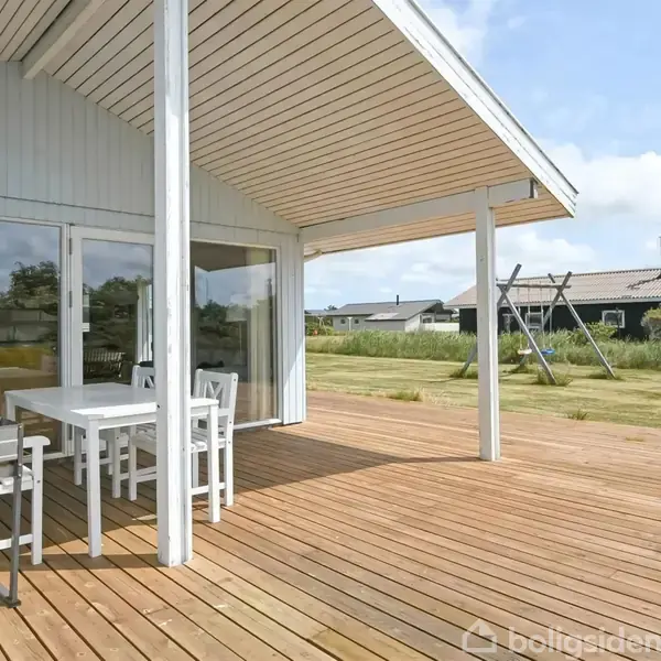 Træterrasse med udendørs spisebord og stole står ved siden af et hvidt sommerhus. I baggrunden ses en græsplæne med gyngestativ og andre huse.
