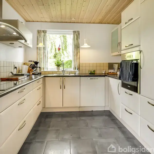 Et moderne køkken med hvide skabe og stålgreb. En ovn er indbygget til højre. På køkkenbordet til venstre ligger køkkenredskaber. I midten ses et vindue med planter udenfor.