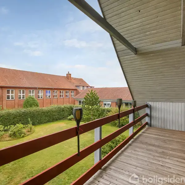 En altan med træplanker og rødt rækværk står stille med udsigt over en velplejet have. I baggrunden ses en bygning med rødt tegltag og flere vinduer.