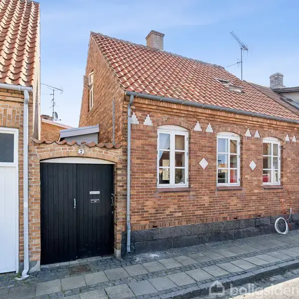 Rødt murstenshus med rødt tegltag, sorte træporte og hvide vinduer. Står langs en brostensbelagt gade i et rækkehusområde. Vejret er klart og solrigt.
