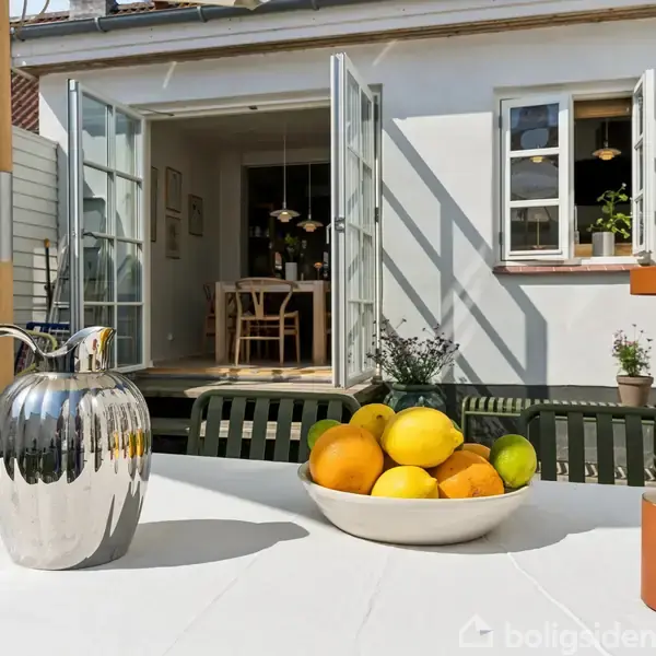 Kande i metal, skål med citrusfrugter og en lampe står på et bord i solrigt gårdmiljø; åbne døre viser ind til en spisestue med træmøbler.