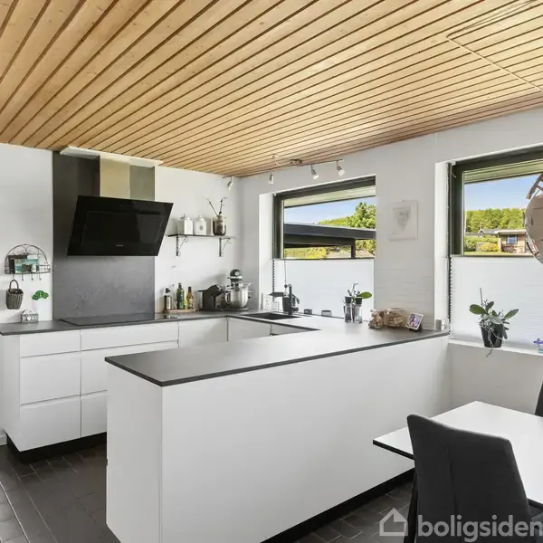 Et moderne køkken med hvide skabe og en sort bordplade. Der er en kogeplade og emhætte. Et spiseområde med stole er ved siden af. Store vinduer giver naturligt lys.