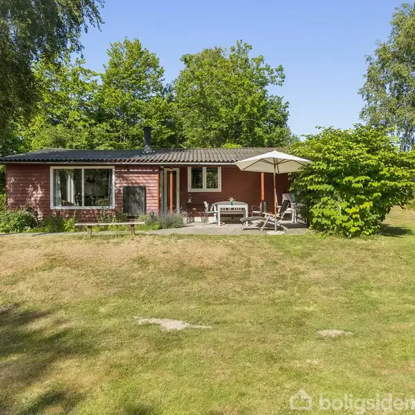 Et rødt træhus med terrasse ligger i en grøn have. Havemøbler og en parasol står på terrassen omgivet af skov og træer.
