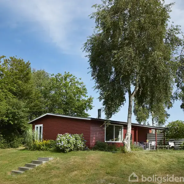 Rødt træhus står stille på en grøn græsplæne med en høj birketræ ved siden af. Omgivet af tæt løv og have med hvidt havebord og stole.