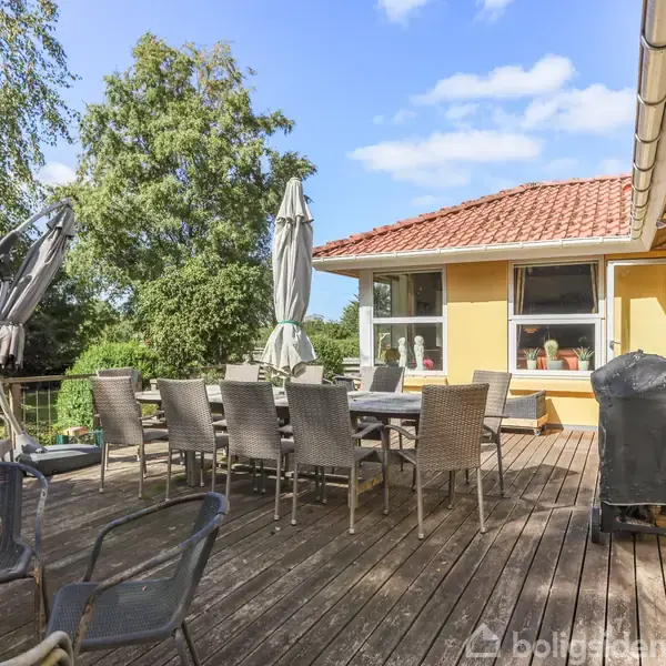 En stor træterrasse med flere havestole og borde under parasoller. En gul bygning med rødt tag bagved, omgivet af grønne træer og en blå himmel.