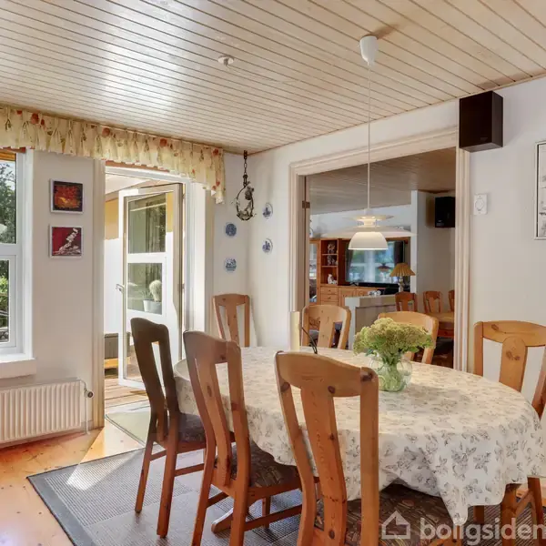 En spisestue med et rundt bord dækket af en hvid dug, omgivet af træstole. Lyse trævægge og loft, store vinduer med udsigt til have.