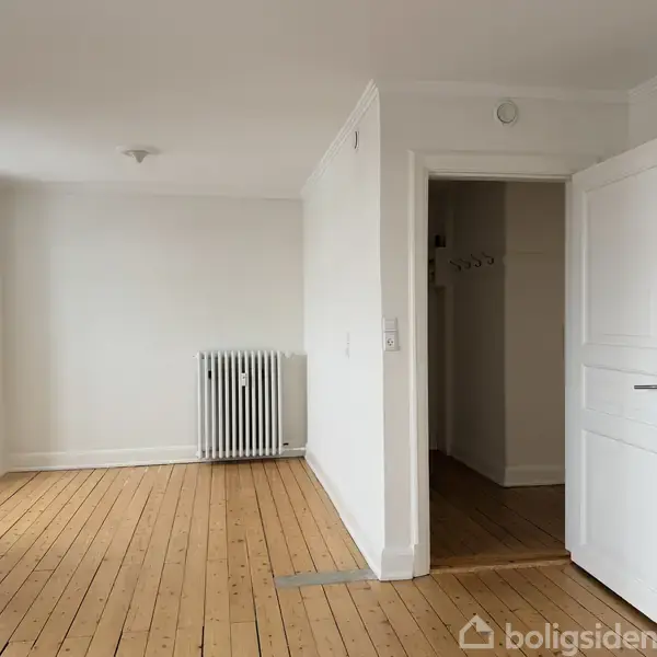 Et tomt rum med trægulv og hvidmalede vægge. En åben dør fører til et andet rum. Der er en radiator ved væggen til venstre.