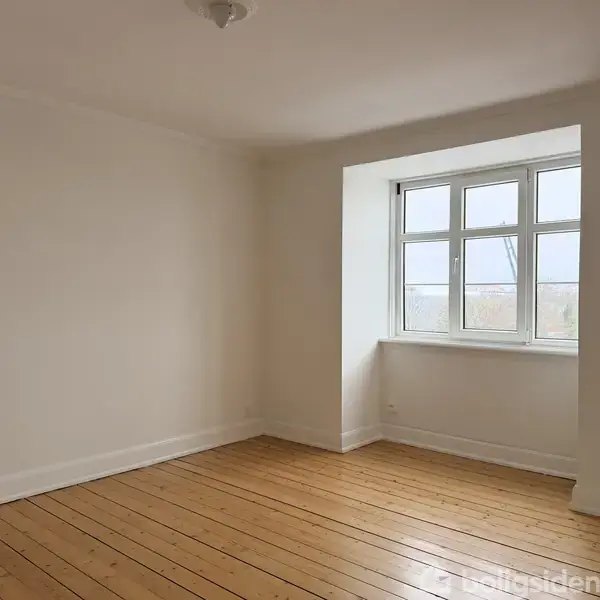 Et tomt rum med lyse trægulve og hvide vægge, domineret af et stort vindue med udsigt til en klar himmel. Rummet er enkelt og minimalistisk indrettet.