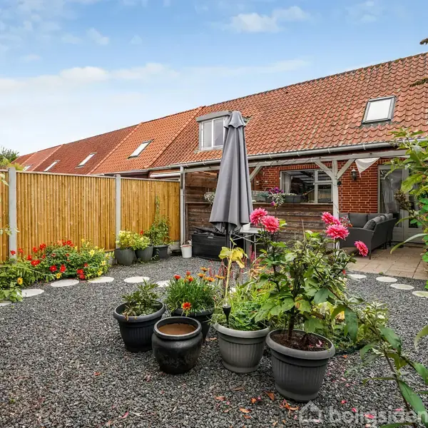 Grusbelagt gårdhave med blomsterkrukker og en parasol. Til venstre er et hegn med beplantning. Baggrunden er et rækkehus med rødt tegltag og terrasse.
