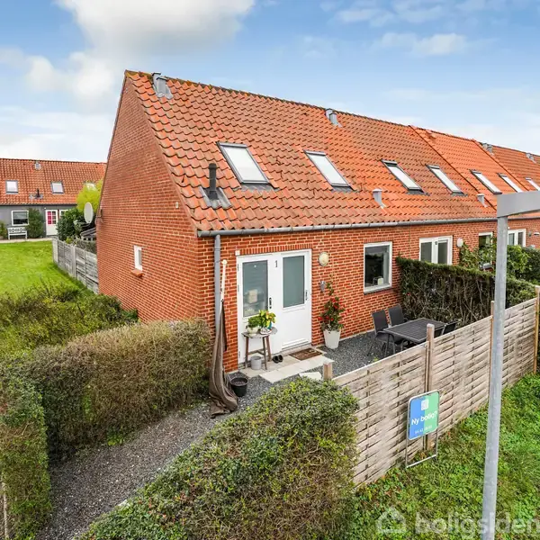 Rækkehus med rødt tegltag og hvide døre, placeret i et boligkvarter med hække og små haver. Ved siden af huset er en flisesti og en lille terrasse med planter.