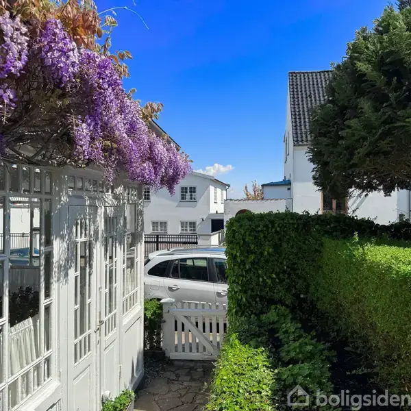 Smuk pergola dækket af lilla blomster over indgang til have. I baggrunden ses en hvid bil parkeret foran hvide huse, omkranset af grønne hække og træer under en klar blå himmel.