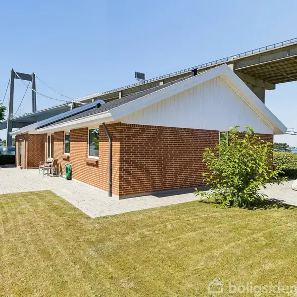Huset er en rød murstensbygning med en flisebelagt terrasse, omgivet af en velplejet have. I baggrunden ses en stor bro over vandet under en klar blå himmel.