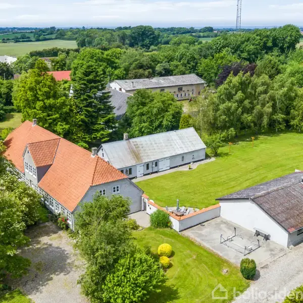 En landlig ejendom med rødt tegltag ses fra oven. Bygningerne er omgivet af grønne græsplæner og træer, beliggende i et åbent, frodigt landskab.