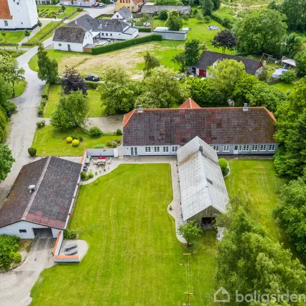 En stor gård med rødt tag ligger omgivet af grønne træer. Gårdspladsen er grusbelagt med en græsplæne i midten. Omgivende huse ses i baggrunden.