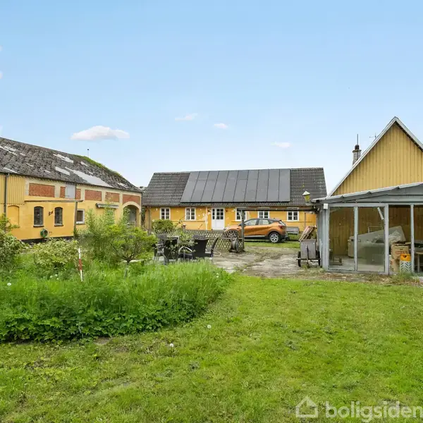 Gårdejerbygninger, gulmalede med sorte tage, står i solskin. Foran en bil parkeret, er der en græsplæne og en terrasse med udemøbler. Omkring ses træer og en klar himmel.