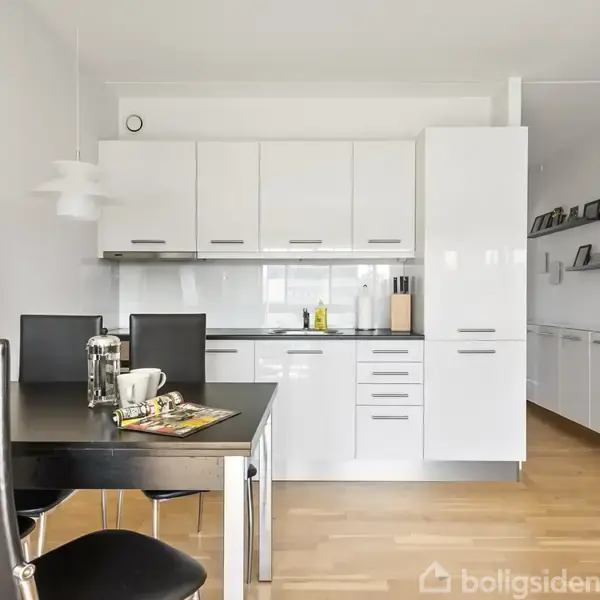 Moderne køkken med hvidt skab og skuffeopstilling. Sorte stole og et bord med kaffekande og magasin. Trægulv og hvidmalede vægge. Enkel indretning med væghylder til højre.