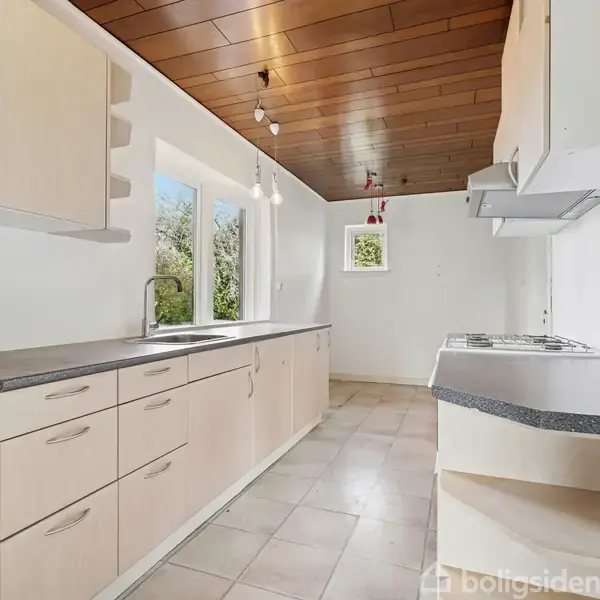 Lyst og rummeligt køkken med lyse træskabe og en lang sort bordplade. Vinduet giver udsigt til grønne omgivelser. Flisegulv og træbeklædt loft med moderne lamper.