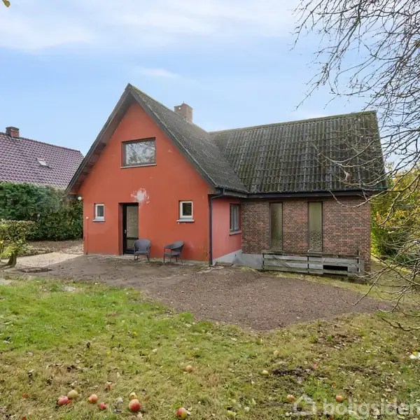 Rødt murstenshus står stille i en have med græs og enkelte nedfaldne æbler. Omkring huset ses buske og et mindre træ, mens en anden bolig er synlig i baggrunden.