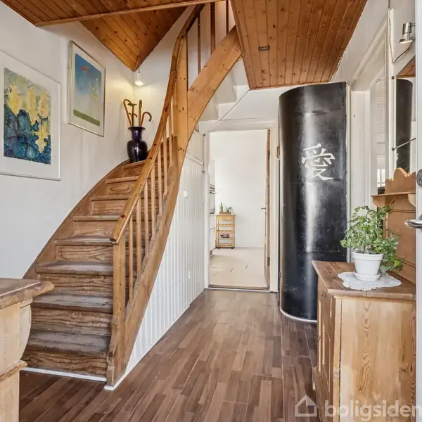 En trætrappe svæver opad i gangen, omgivet af hvide vægge og kunstværker. En sort cylinder med asiatisk tegn står oprejst; ved siden af står en plante på en kommode.