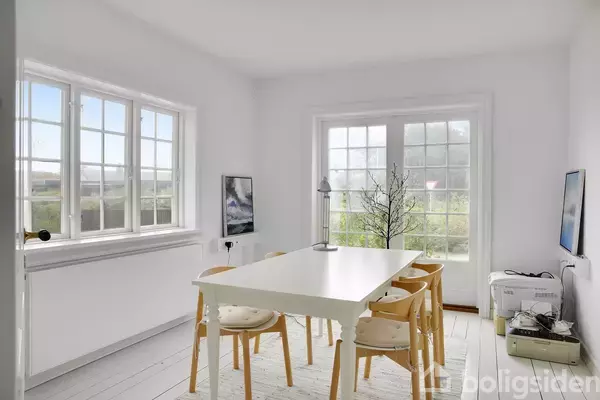 Hvidt spisebord med seks træstole står i lys, rummelig spisestue. Store vinduer og glasdør slipper naturligt lys ind, med udsigt til grøn have. Enkel indretning med minimalistisk stil.