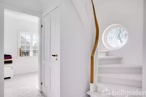 En hvid, snoet trappe med trægelænder står ved et rundt vindue. I baggrunden ses et lyst rum med vindue, der lukker lys ind.