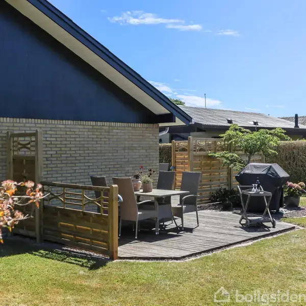 Husets terrasse har havemøbler og grill til afslapning. Omgivet af en velplejet græsplæne og buske. Klimavenligt design med delvis solbelysning under klar himmel.