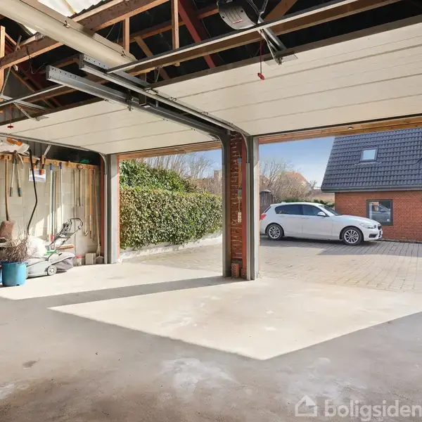 Garage med åbne porte, med udsigt til en indkørsel med en parkeret bil. Indenfor ses cykel, værktøj og opbevaringskasser, under et tag med synlige bjælker.