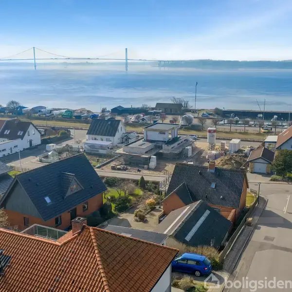 Byhuse ved kysten med udsigt over en bred bro og stille vand i baggrunden. Solskin oplyser husenes tage og den omkringliggende vej.
