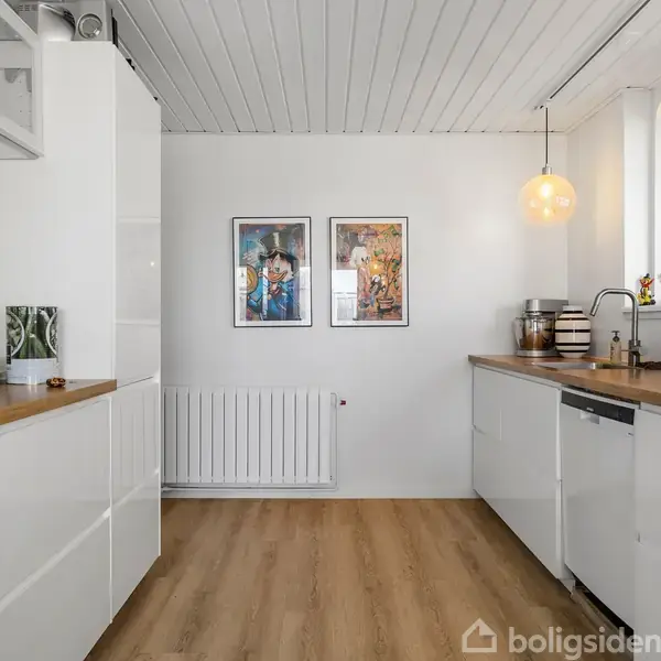 Et lyst køkken med hvide skabe og træbordplader. To billeder hænger på væggen. Vinduerne til højre lader naturligt lys komme ind. To loftlamper hænger over bordpladen.