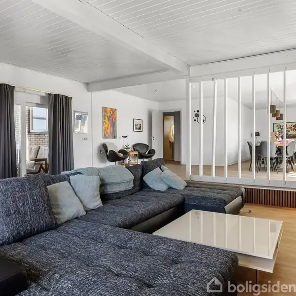 En stor, mørkegrå sofa står i stuen med bløde puder. Den er placeret foran et lavt, hvidt sofabord. Et åbent rum er bagved, delt af hvide stolper.