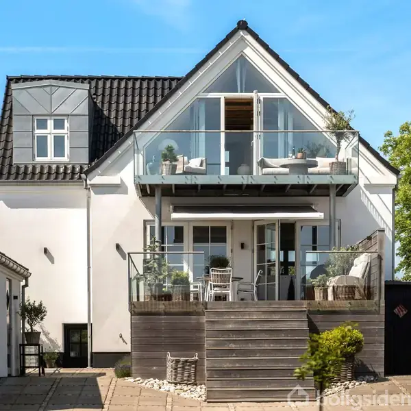 Et hvidt hus med sort tag har en udendørs terrasse med glasrækværk, møbler og planter. Det er omgivet af frodige træer under en klar blå himmel.