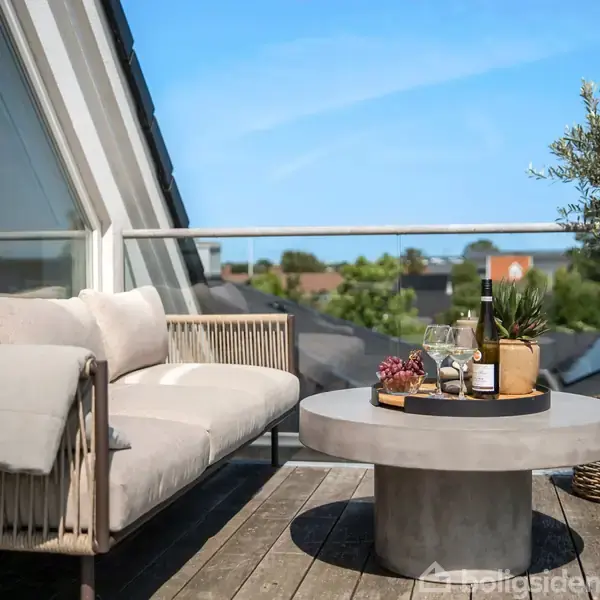 Sofa på altan: En lys sofa står på en udendørs træterrasse ved siden af et rundt betonbord med vin og glas, omgivet af grønne planter under en klar blå himmel.